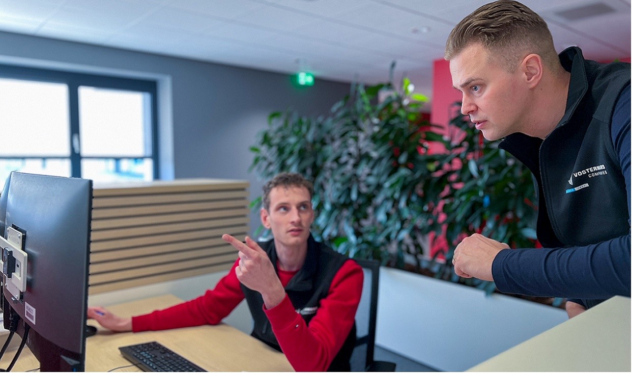 Twee mannen in een kantooromgeving bespreken iets op een computerscherm, waarbij één man zit en wijst naar het scherm terwijl de andere man staat en aandachtig kijkt.