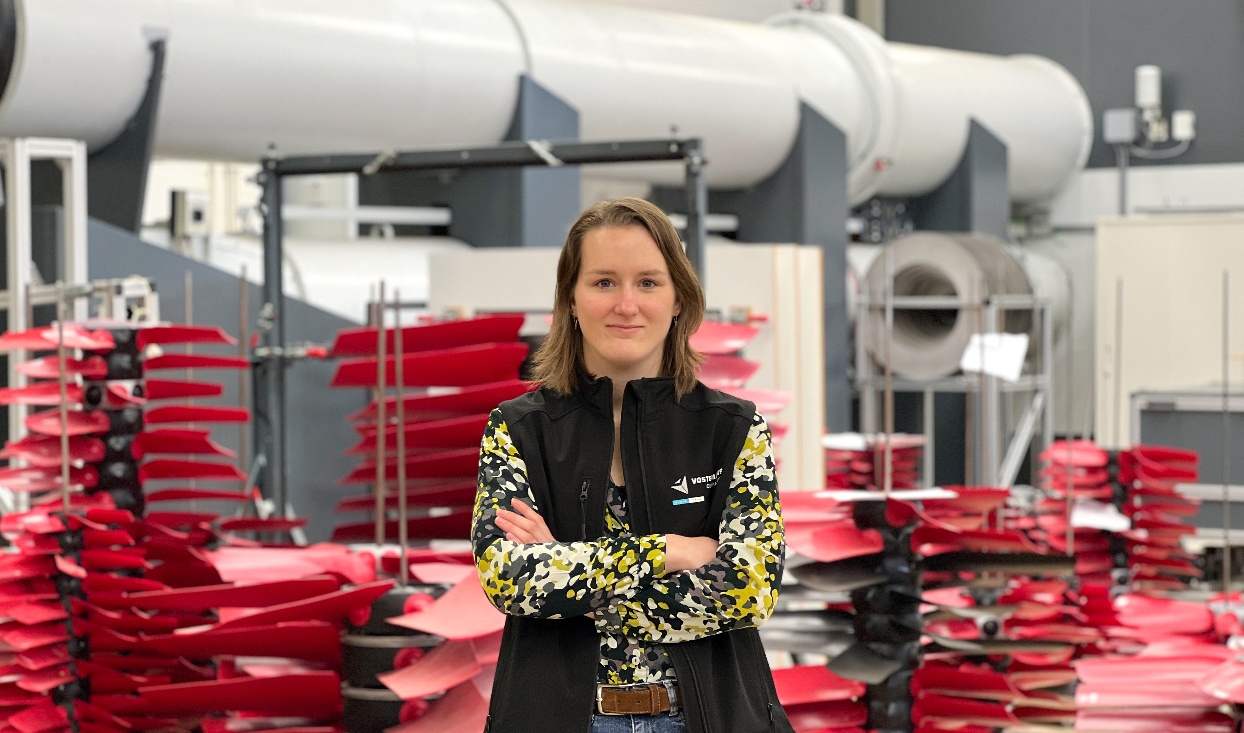 Frau steht mit verschränkten Armen in einer Fabrikhalle, umgeben von roten Teilen und Industrieanlagen im Hintergrund.