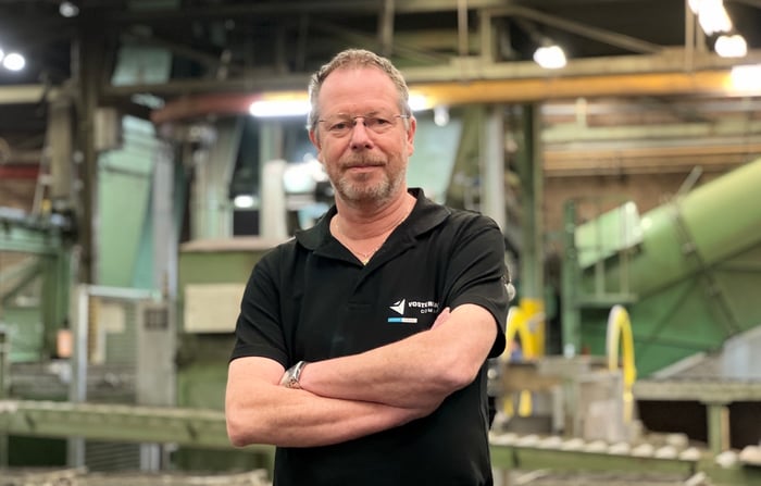 Man with glasses and beard standing confidently with arms crossed in an industrial setting, wearing a black polo shirt with 'Voith Hydro' logo, machinery and equipment visible in the background.