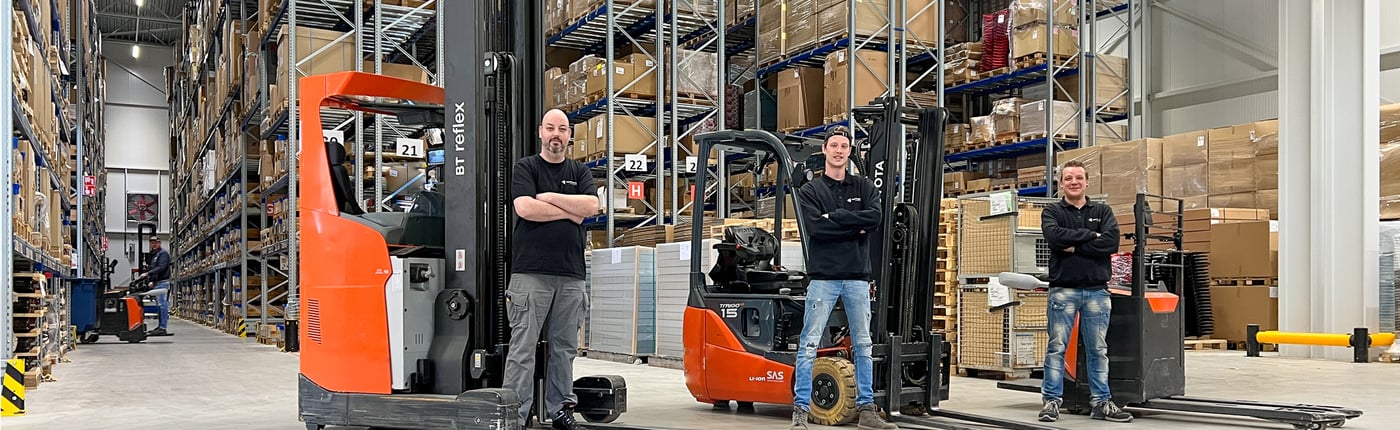 Three men are standing proudly in a large warehouse surrounded by tall racks filled with inventory. They are positioned next to forklifts, ready for operations.