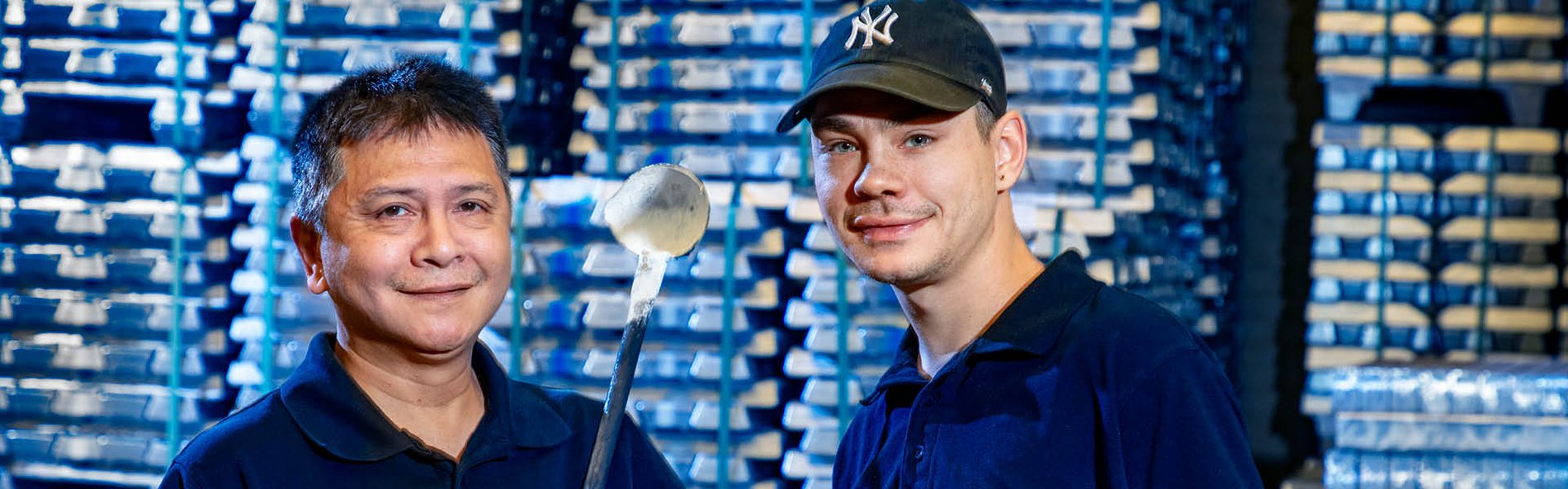 Twee mannen glimlachen naar de camera in een fabrieksomgeving. Een van hen houdt een metalen lepel vast, en op de achtergrond zijn gestapelde metalen blokken te zien, wat een industriële sfeer creëert.