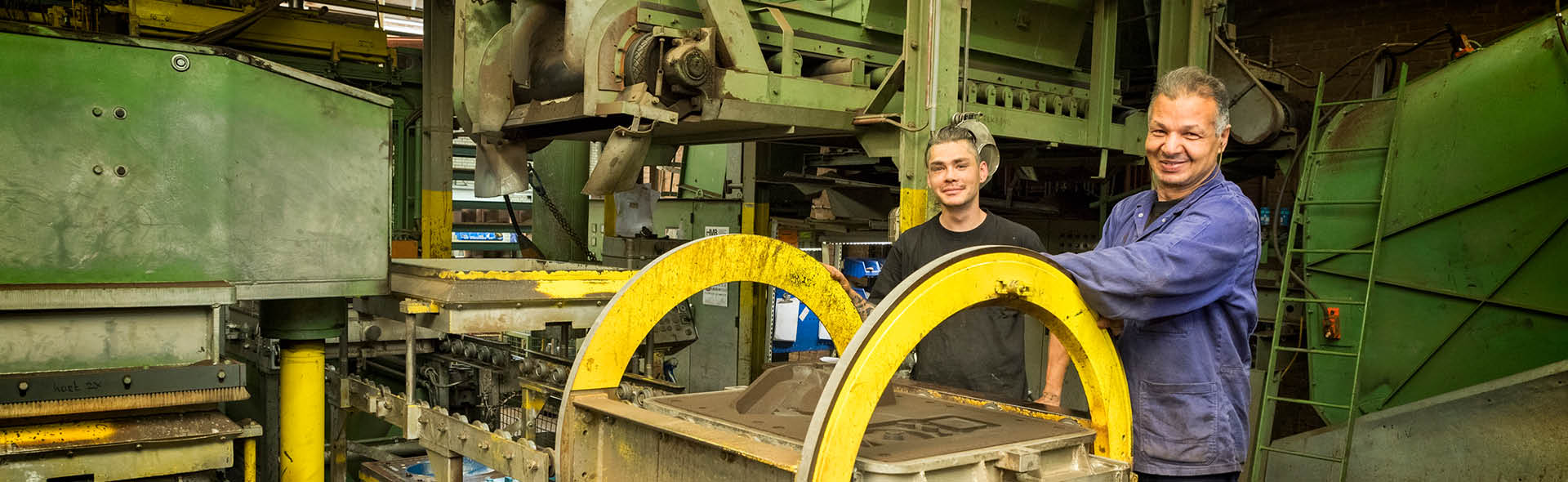 Deux hommes se tiennent dans une fonderie, souriant à la caméra. Ils sont entourés de grandes machines industrielles vertes et jaunes utilisées pour le moulage en sable.