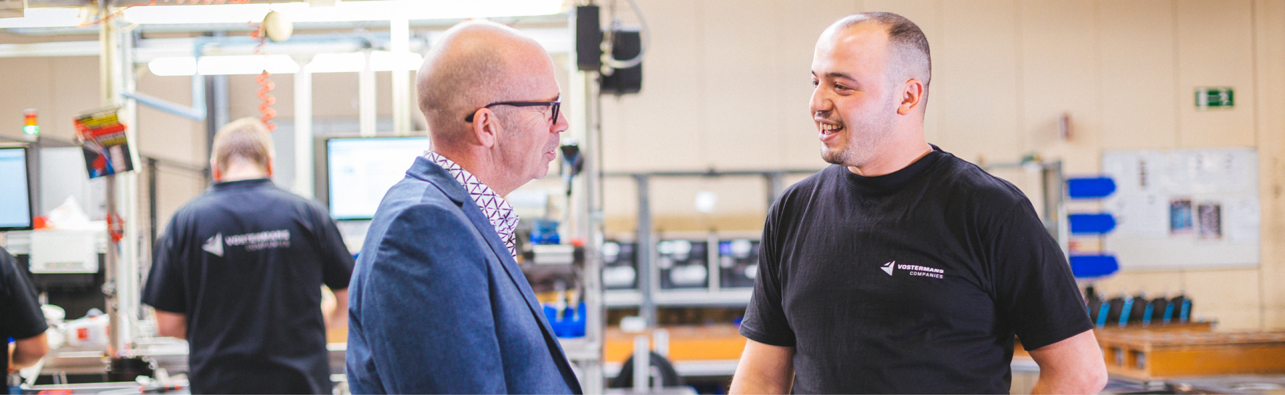 Two men are engaged in conversation in a factory setting. One man is wearing glasses and a suit, while the other is wearing a black shirt with a company logo. Other employees are working in the background.