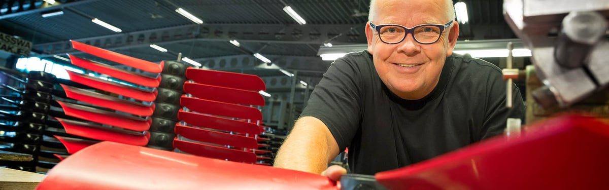 Een man glimlacht naar de camera terwijl hij over een rood industrieel onderdeel in een fabrieksomgeving leunt. Verschillende soortgelijke rode onderdelen zijn netjes op de achtergrond gestapeld.