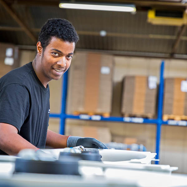 Een man met werkhandschoenen glimlacht naar de camera terwijl hij in een fabriek werkt, omringd door grote onderdelen en met opgeslagen materialen op de achtergrond.