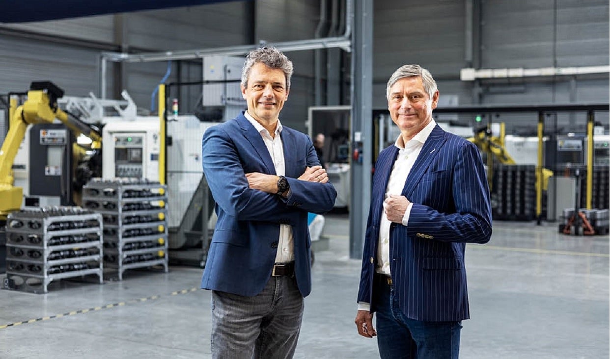 Deux hommes en tenue de travail se tiennent dans un hall d'usine moderne, entourés de machines automatisées et d'équipements industriels, et regardent la caméra avec assurance.