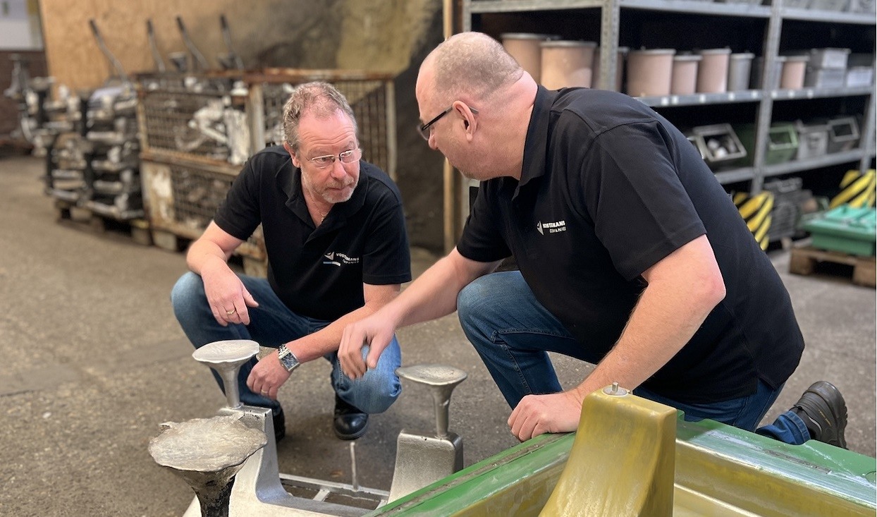 Twee mannen in werkkleding bespreken een metalen onderdeel in een industriële omgeving, waarbij ze gehurkt bij het object zitten en omringd zijn door gereedschap en machines.