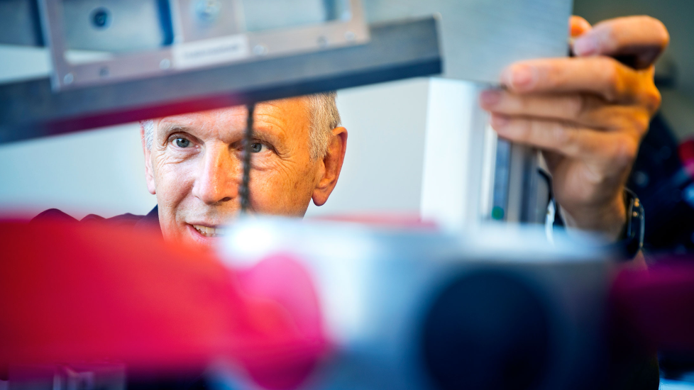 An older man is carefully examining a technical machine or instrument, with his face partially visible between the components. His expression is focused and curious, indicating a deep engagement with his work.