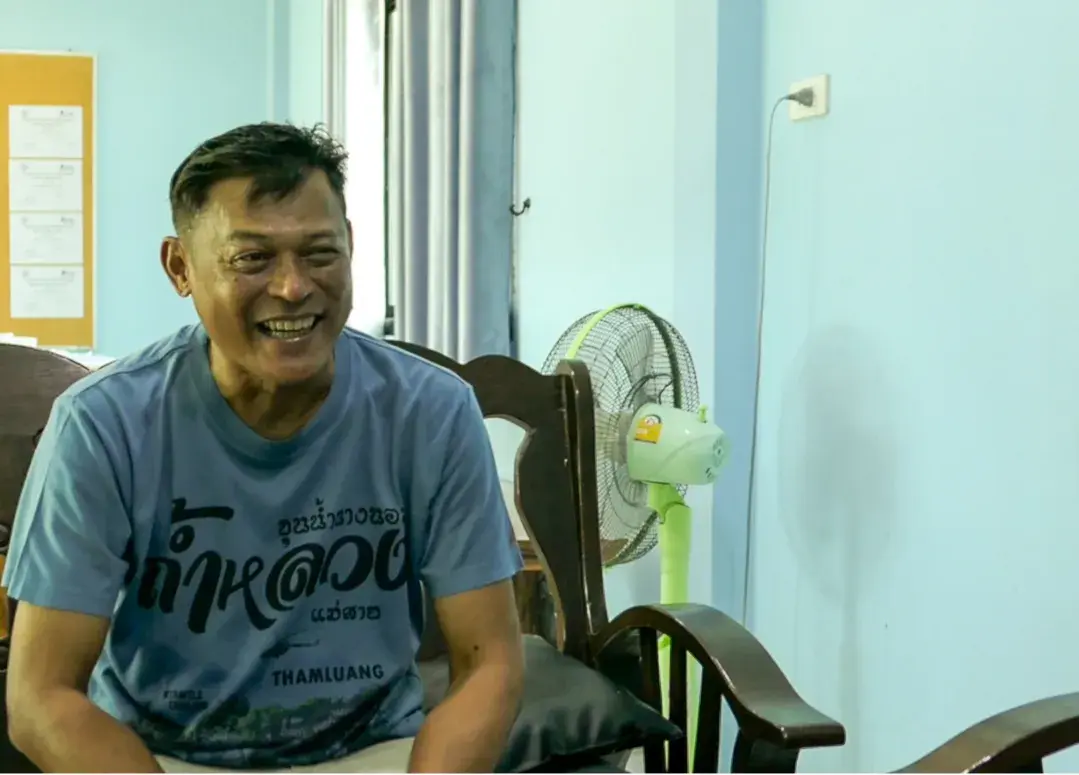 Smiling man sitting on a chair with a fan in the background