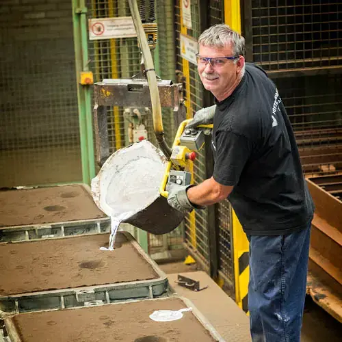 Worker smiling while pouring molten metal into sand casting mold in a factory