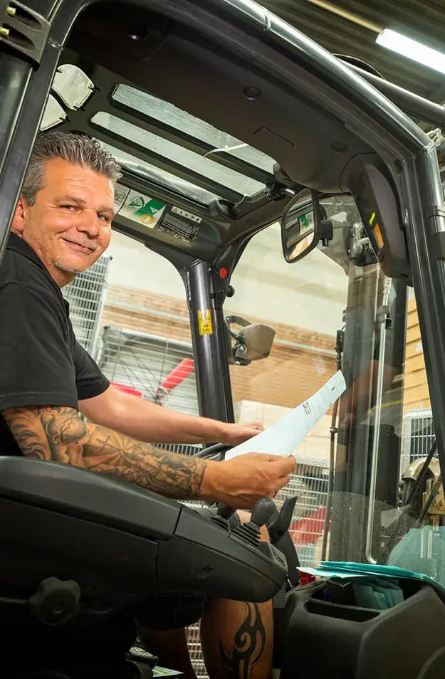 Warehouse employee sitting in a forklift, holding documents and smiling at the camera