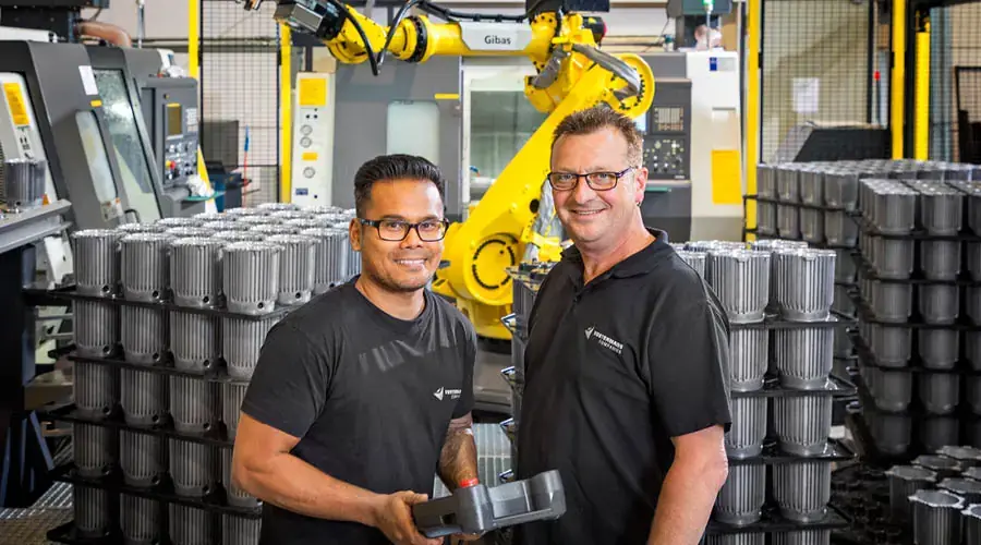 Twee medewerkers van de metaalonderdelenfabriek glimlachen, omringd door stapels metalen onderdelen en machines