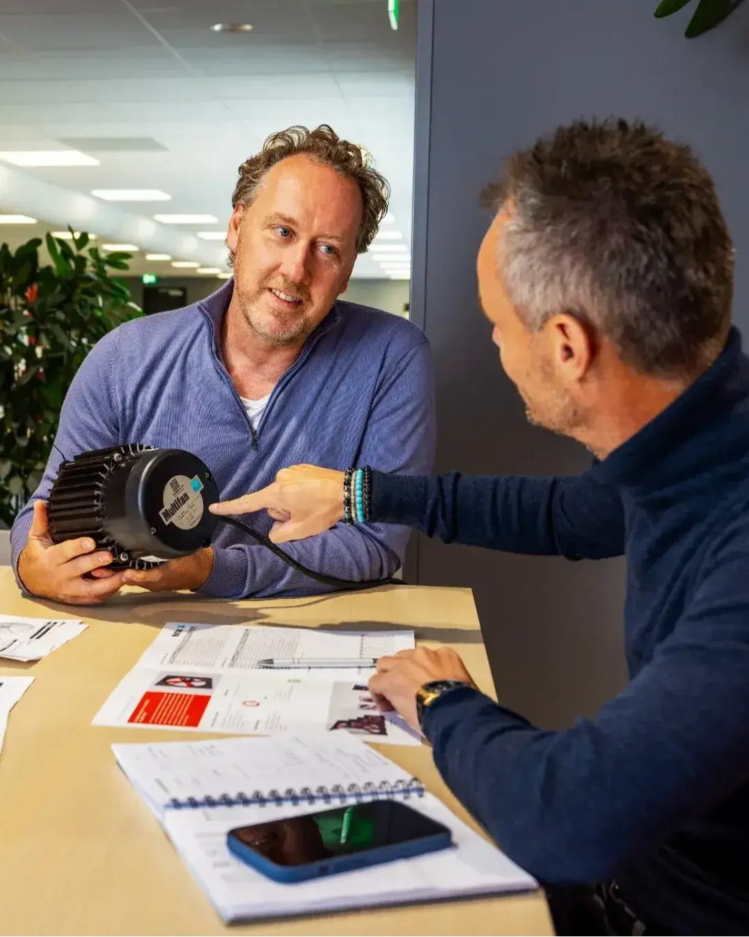 Drei Personen besprechen technische Dokumente und halten ein Motorbauteil in einer Bürobesprechung