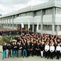 Una gran foto de grupo de empleados reunidos frente a un moderno edificio celebrando el 50 aniversario de la empresa con banderas y sonrisas