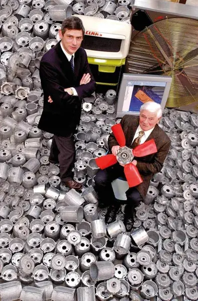 Deux hommes posent au milieu d'une pile de composants métalliques l'un tenant un ventilateur et l'autre se tenant avec assurance