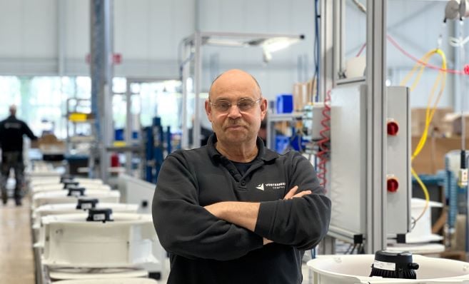 A man is standing in a bright and modern factory setting, with his arms crossed and wearing a black company shirt. Behind him are assembly line components and various pieces of machinery.