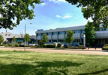 Exterior del edificio de Vostermans Ventilation en un día soleado, rodeado de árboles y hierba verde.